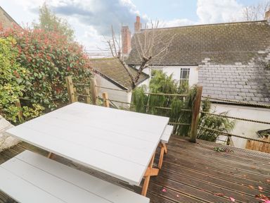 An outdoor wooden deck with a white picnic table and benches surrounded by plants and trees at Belmont in Newlyn