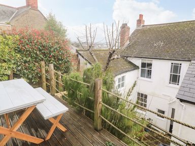 A wooden deck with a white table and bench overlooking a white building and garden at Belmont in Newlyn