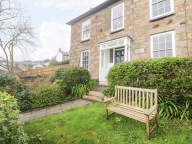 A garden with a wooden bench and shrubs in front of a stone house with white windows and a green door at Belmont in Newlyn