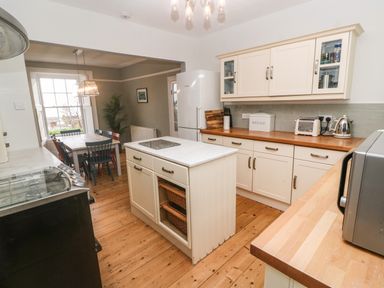 A kitchen with a central island wooden flooring white cabinets and an adjacent dining area with a wooden table and chairs at Belmont in Newlyn