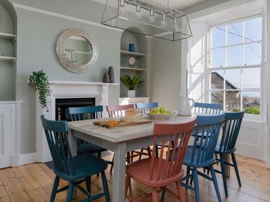 A dining room with a wooden table surrounded by colorful chairs and a window showing rooftops at Belmont in Newlyn