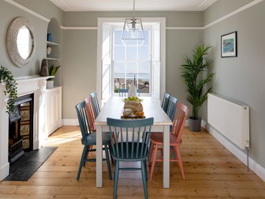 A dining room with wooden floor a white table blue and pink chairs a fireplace and a large window with a sea view at Belmont in Newlyn