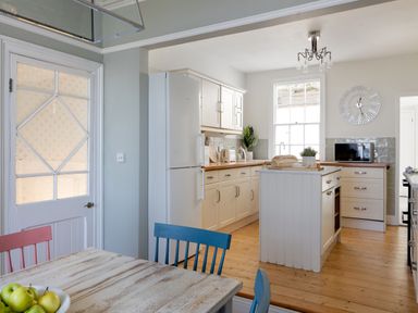 A kitchen with white cabinets a wooden island wooden floor and a dining table with colored chairs at Belmont in Newlyn