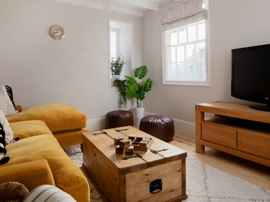 A living room with a yellow sofa wooden coffee table two brown poufs a television on a wooden stand and plants at Belmont in Newlyn
