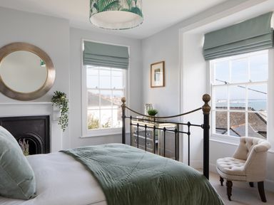 A bedroom with a bed a round mirror a dresser an upholstered chair and two windows at Belmont in Newlyn
