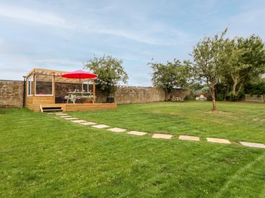 A backyard with a wooden deck with a red umbrella picnic table and string lights a stone pathway leading to the deck green grass trees and a brick wall at The Vault House in Bellingham