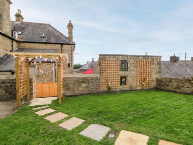 A grassy garden with a stone pathway leading to a wooden pergola and stone walls with wooden trellises at The Vault House in Bellingham