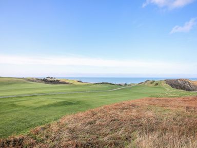 A grassy landscape with a golf course and a road leading towards buildings near the coast at Min Y Mor - Morfa Nefyn