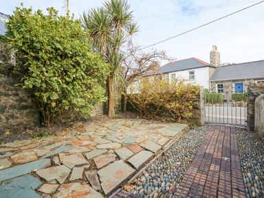 A stone paved patio area with shrubs and a palm tree near a gated entrance and stone wall at Min Y Mor - Morfa Nefyn