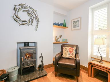 A corner with a wood burning stove a leather armchair with a patterned cushion beside a window with shutters and a small wooden lamp table at Min Y Mor - Morfa Nefyn