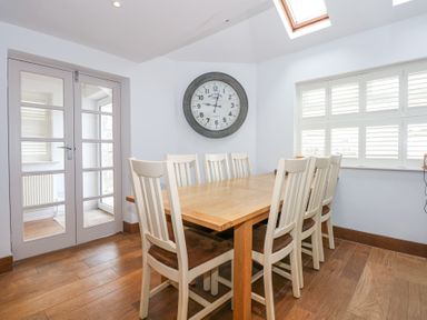 A dining room with a wooden table and eight chairs a large wall clock and a window with shutters at Min Y Mor - Morfa Nefyn
