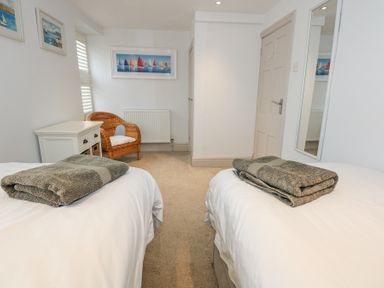 A bedroom with two beds with towels on them a wicker chair a small white chest of drawers a wall mirror and framed pictures on the wall at Min Y Mor - Morfa Nefyn