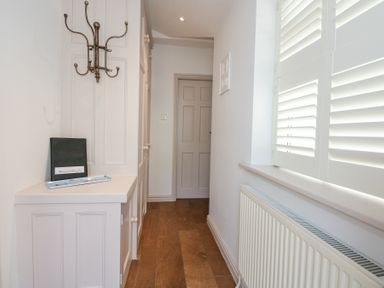 A narrow hallway with wooden floor a closed door white cabinets a wall-mounted coat rack and a radiator with window shutters at Min Y Mor - Morfa Nefyn