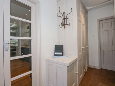 A hallway with white built-in cabinets a corner shelf with a folder and tray a wall-mounted coat rack and a glass-paneled door showing a kitchen at Min Y Mor - Morfa Nefyn