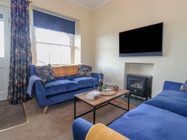A living room with blue sofas a wooden coffee table a wall mounted tv and a small stove at Primrose Cottage in Ambleside