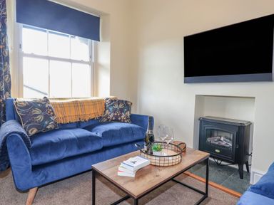 A living room with a blue sofa two patterned cushions a wooden coffee table with a tray and books a television mounted above a fireplace at Primrose Cottage in Ambleside
