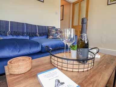 A living room with a blue sofa a wooden coffee table with a basket a book a bottle of prosecco and two wine glasses at Primrose Cottage in Ambleside