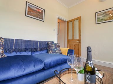 A living room with a blue couch, two wine glasses, a bottle of prosecco on a wooden table, wooden door and framed pictures on walls at Primrose Cottage in Ambleside