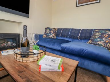 A wooden coffee table with a bottle of prosecco two glasses and books in front of a blue sofa with patterned cushions at Primrose Cottage in Ambleside