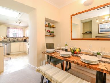 A dining area with a wooden table set with plates and wine glasses a bench and chairs and a kitchen with appliances visible through an open doorway at Primrose Cottage in Ambleside