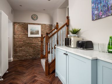 A kitchen area with light blue cabinets a microwave a toaster glasses and a bottle next to wooden stairs and an exposed brick wall with a clock and a painting at Owl Cottage in Morpeth