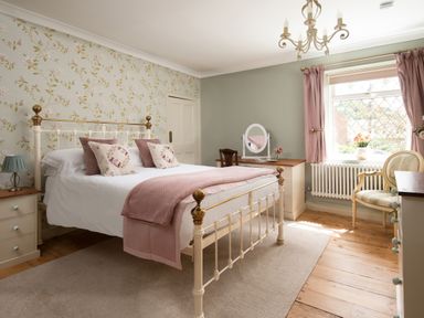 A bedroom with a white and gold bed with pink pillows and throw a floral wallpaper on one wall a window with pink curtains a chair a rug and a vanity desk at Owl Cottage in Morpeth