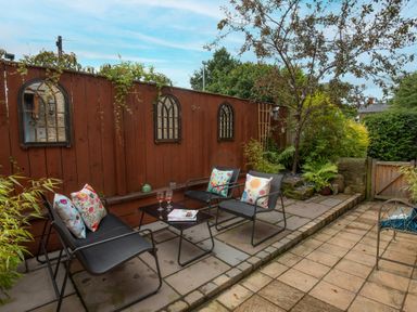 A patio area with black metal chairs and a table with drinks and pillows against a wooden fence with plants and a tree at Owl Cottage in Morpeth
