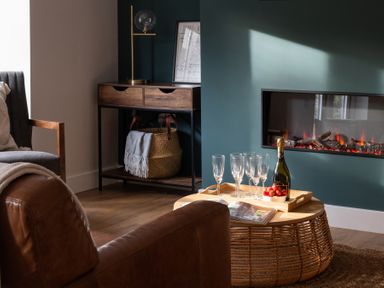 A living room with a leather chair a wooden cabinet a basket a fireplace and a wicker coffee table with champagne glasses strawberries and an open magazine at Hebog in Beddgelert
