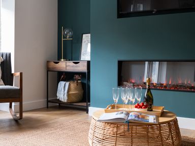 A living room corner with a wicker coffee table holding glasses strawberries and a bottle a fireplace and a wooden side table with a lamp and basket at Hebog in Beddgelert