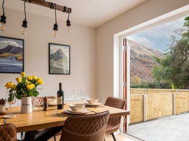 A dining room with a wooden table set with bowls wine bottle and flowers next to an open door showing a mountain view at Hebog in Beddgelert