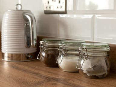 Three glass jars with tea bags sugar and coffee next to an electric kettle on a wooden kitchen counter