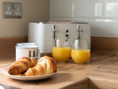 A kitchen countertop with two croissants on a plate two mugs with tree designs two glasses of orange juice and a toaster at Hebog in Beddgelert
