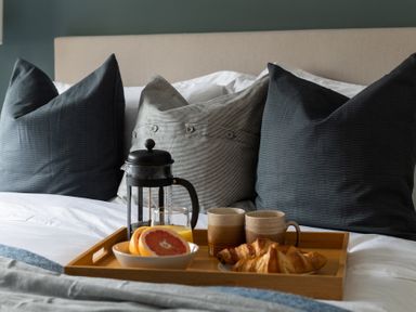 A wooden tray on a bed with a glass French press, orange juice, coffee mugs, croissants, and sliced grapefruit with pillows in the background at Hebog in Beddgelert