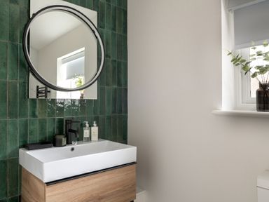 A bathroom sink with a wooden cabinet green tiled wall round mirror and a window with a plant at Hebog in Beddgelert