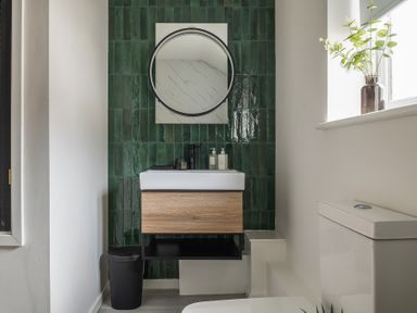 A bathroom with a sink, mirror, green tiled wall, trash bin, toilet, and a window with a plant at Hebog in Beddgelert