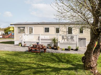 A beige mobile home with a white railing and outdoor furniture surrounded by potted plants and grass at The Maples Steel Green near Haverigg