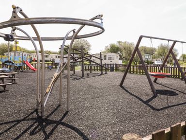 An outdoor playground with climbing frames a swing and a slide with trailers and trees in the background at The Maples in Steel Green near Haverigg