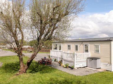 A beige mobile home with white railing and potted plants beside a tree and picnic table at The Maples in Steel Green near Haverigg