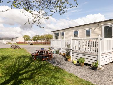 An outdoor area with a white deck attached to a mobile home and wooden picnic table on grass at The Maples in Steel Green near Haverigg