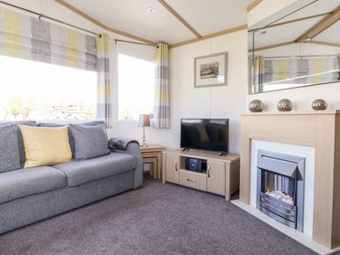 A living room with a gray sofa a television on a wooden stand a fireplace and windows with striped curtains at The Maples in Steel Green near Haverigg