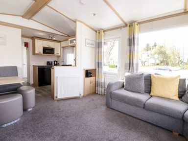 A living room with a grey couch and yellow pillows and a small kitchen area with a microwave and a fridge at The Maples in Steel Green near Haverigg