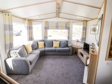 A living room with a grey sectional sofa yellow and grey cushions a television on a wooden stand and striped curtains at The Maples in Steel Green near Haverigg