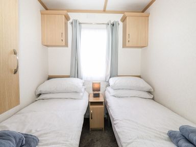 A bedroom with two single beds separated by a small wooden nightstand with a lamp and blue curtains at The Maples in Steel Green near Haverigg