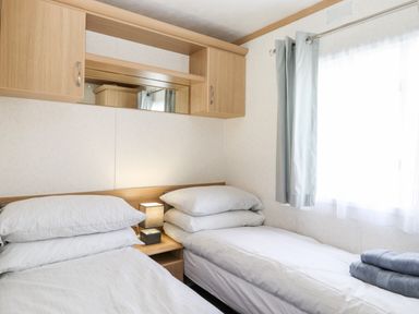 A bedroom with two single beds a bedside table with a lamp overhead cabinets and a window with curtains at The Maples in Steel Green near Haverigg