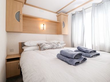 A bedroom with a bed with white bedding and two folded towels on it next to light wood cabinets and a window with curtains at The Maples Steel Green near Haverigg