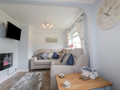 A living room with a gray sofa and cushions a wall-mounted tv an electric fireplace and a wooden side table at Merryfield Lodge in Rosecraddoc near Liskeard