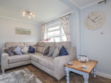 A living room with a beige sectional sofa with cushions a wooden side table with decor under a large wall clock at Merryfield Lodge Rosecraddoc near Liskeard