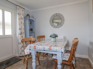 A dining area with a table covered with a lighthouse patterned cloth and wooden chairs around it with a round mirror on the wall and a blue bookshelf in the corner at Merryfield Lodge Rosecraddoc near Liskeard