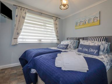 A bedroom with two single beds with blue bedding and pillows next to a window with curtains and a wall-mounted TV at Merryfield Lodge Rosecraddoc near Liskeard