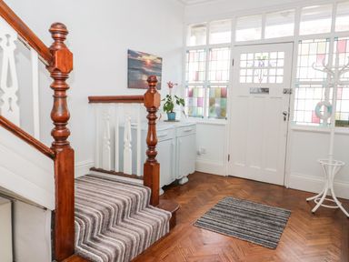 An entryway with a wooden staircase, a striped carpet runner, a small cabinet with a plant, stained glass windows, and a coat rack at 3 St Davids Place in Llandudno